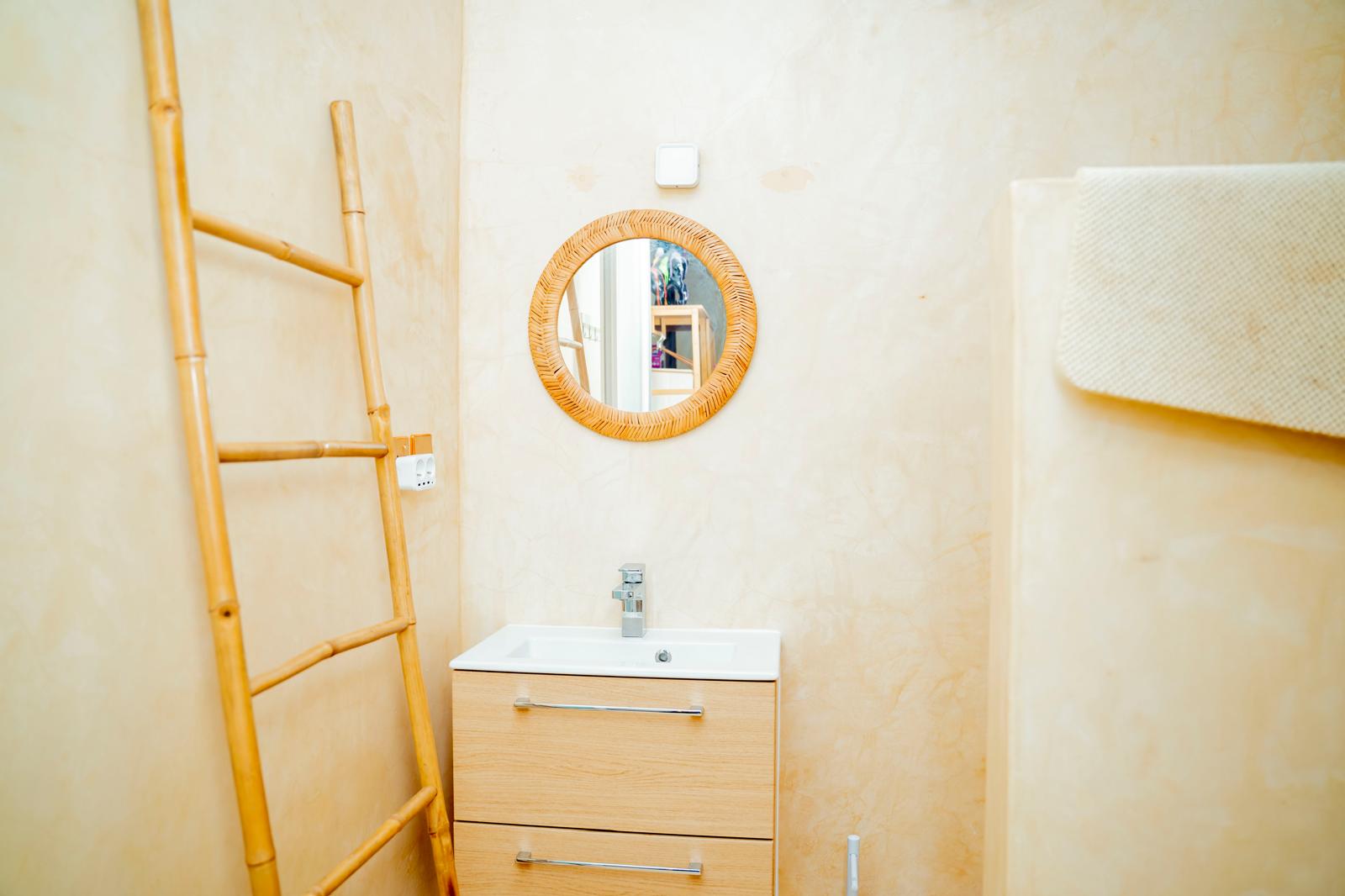 Salle d'eau de la villa en enduit à la chaux, miroir rond en rotin et vasque en bois à Zanzibar
